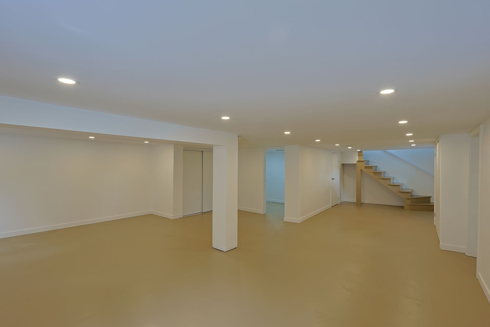 Empty unfinished basement with white walls, beige floor, recessed lights, and stairs leading up on the right