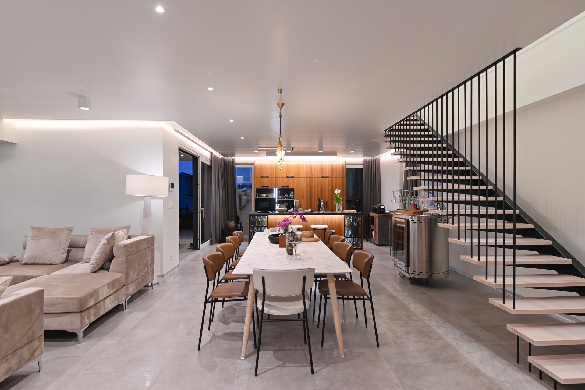 Modern open-plan dining and living room with staircase, gray sofa, and warm kitchen lighting