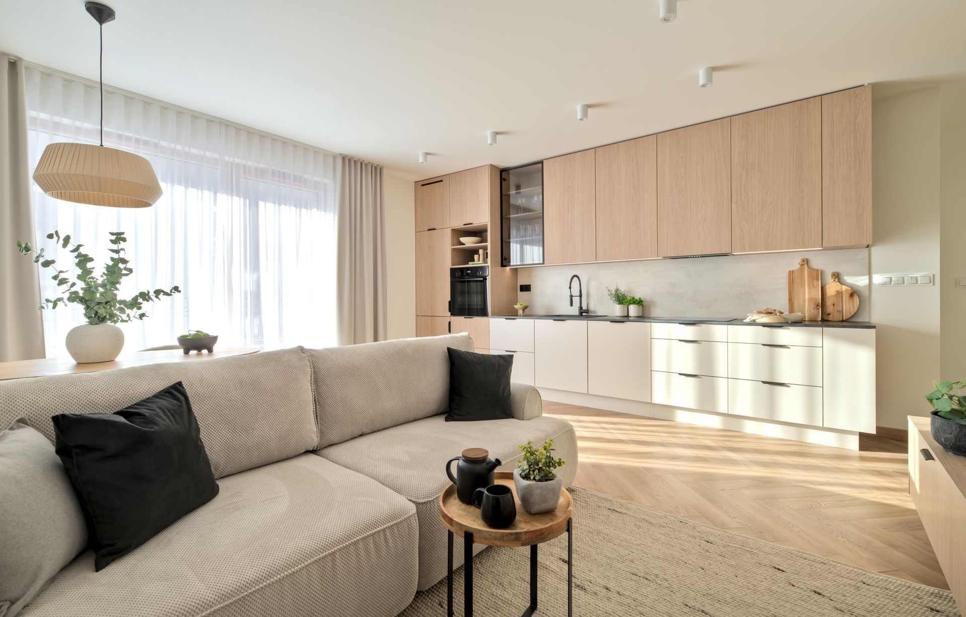 Bright modern living room and kitchen with beige sofa, wood cabinets, and large windows
