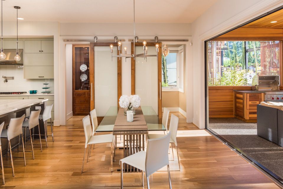 Modern dining room with long table, white chairs, wooden floor, and open kitchen leading to a patio.