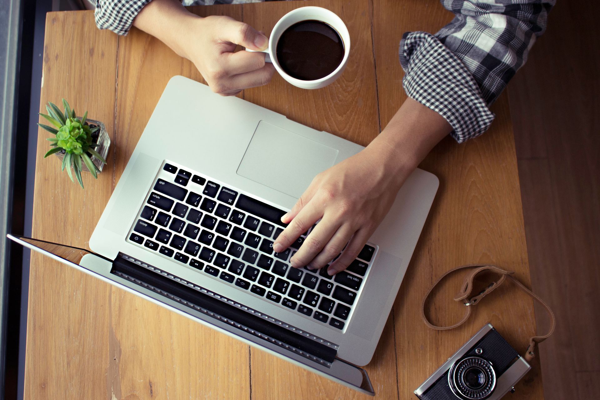 Hands typing on a laptop at a wooden desk with coffee, a plant, and a camera.