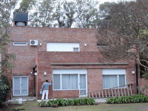 Una persona subida a una escalera pintando el exterior de una casa de ladrillo, con árboles al fondo