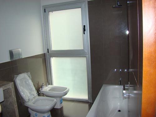 Baño pequeño con inodoro, lavabo, ventana esmerilada y bañera con mampara de ducha de cristal.