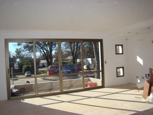 Luminoso salón vacío con puertas correderas de cristal, suelo bañado por el sol y vistas a los árboles del exterior.
