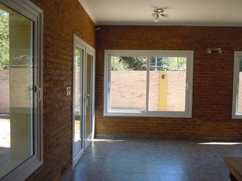 Habitación vacía con paredes de ladrillo, grandes ventanales y una puerta de cristal, iluminada por la luz natural