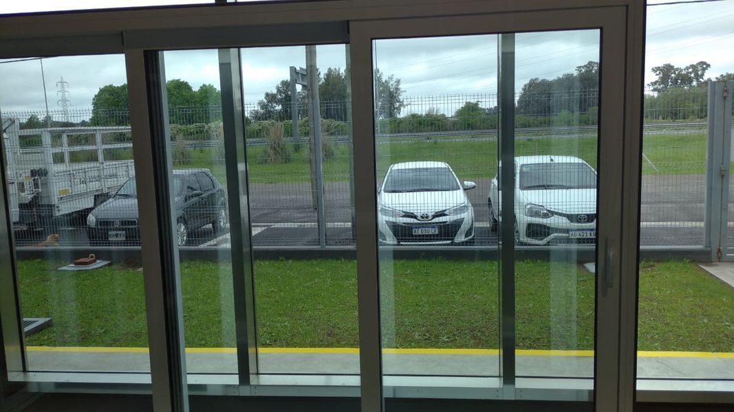 Vista a través de grandes ventanales de coches aparcados en un terreno con césped bajo un cielo nublado