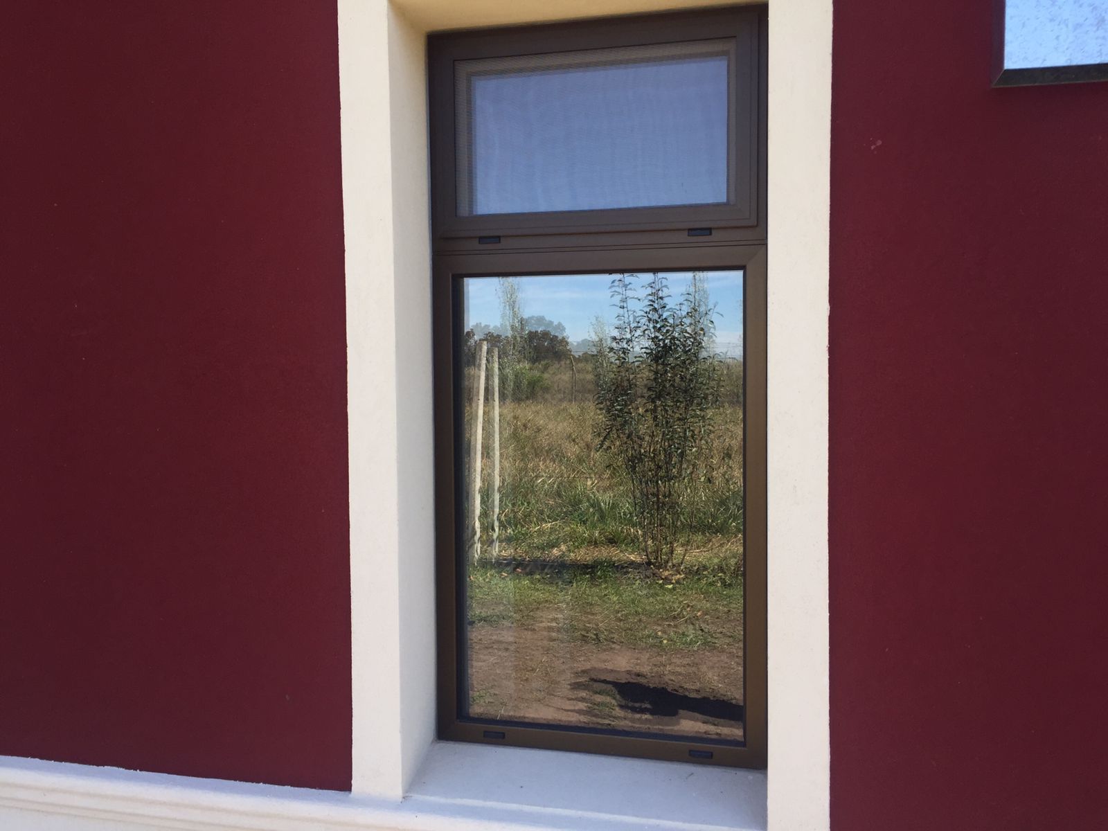 Ventana alta con marco oscuro en una pared burdeos, con vistas a un campo cubierto de hierba.