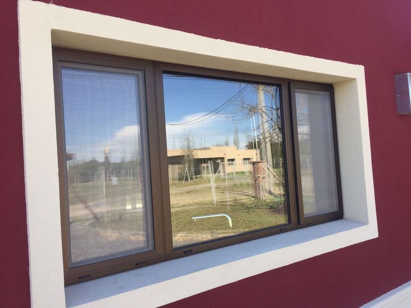 Pequeña ventana con marco blanco en una pared exterior roja, que refleja un paisaje cubierto de hierba y varios edificios.