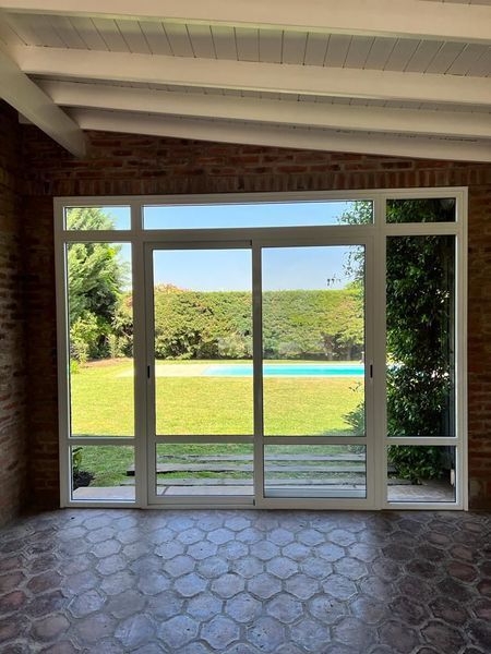 Patio vacío con vistas a un césped soleado, una piscina azul y árboles al otro lado de una gran puerta corrediza de vidrio