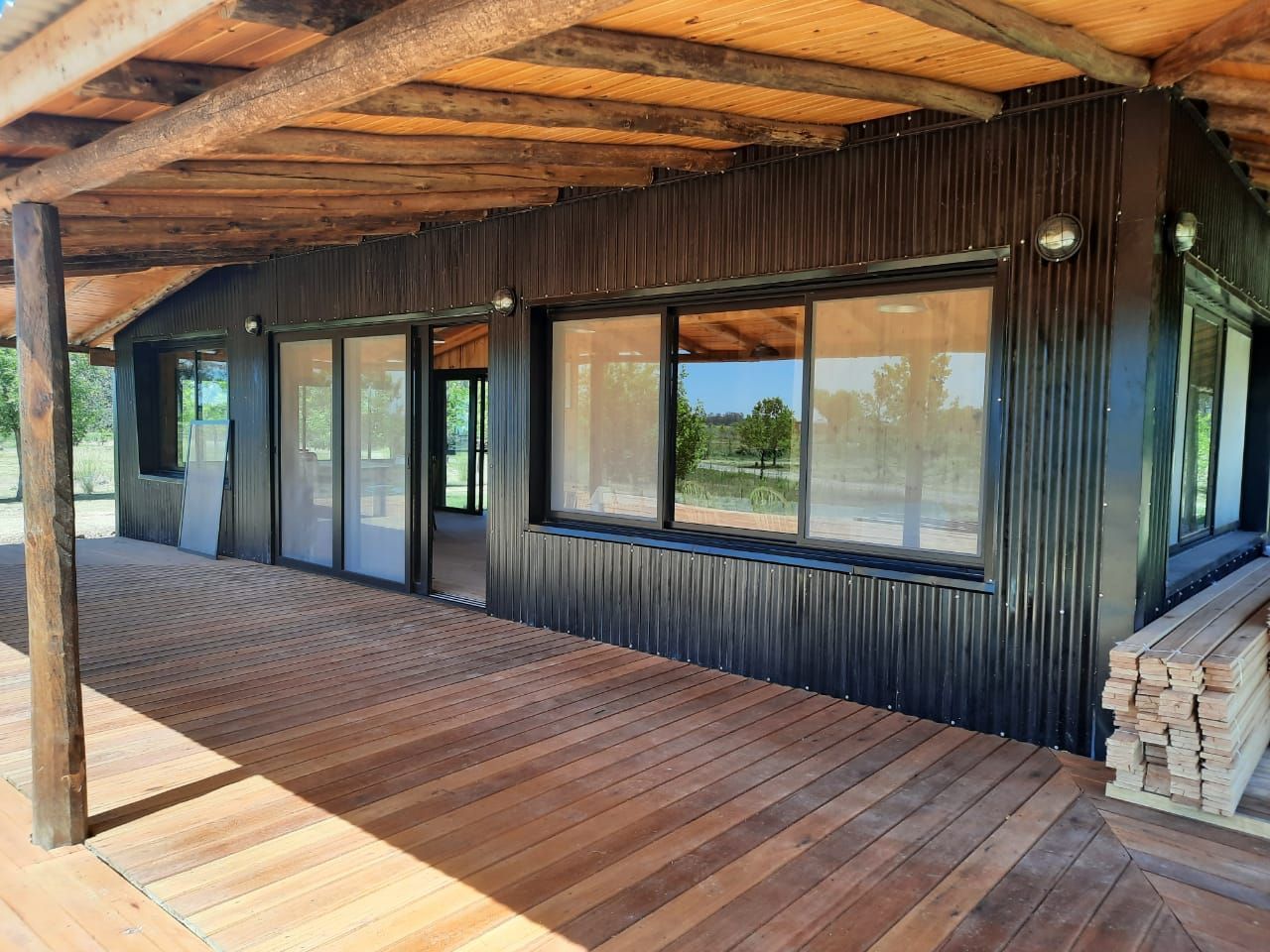 Porche cubierto de madera con revestimiento negro, grandes ventanales y columnas de piedra apilada.