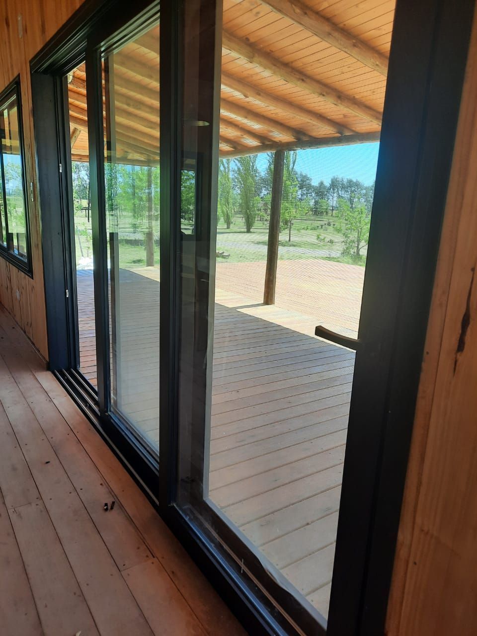 Terraza de madera junto a una cabaña con paredes de cristal, con vistas a un patio de arena y árboles bajo un techo cubierto.