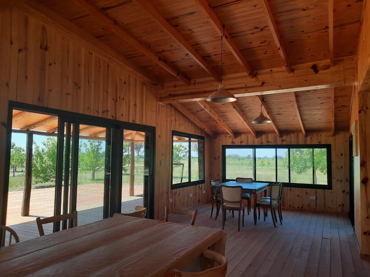 Interior de una cabaña de madera con grandes ventanales, una mesa de comedor y sillas con vistas a un paisaje espectacular al aire libre