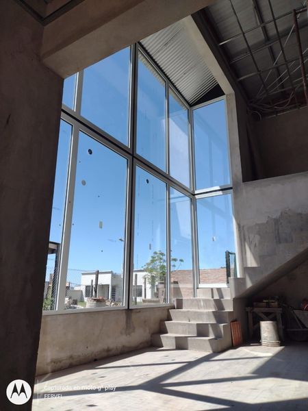 Interior moderno sin terminar, con ventanales del suelo al techo, escaleras de hormigón y abundante luz solar.