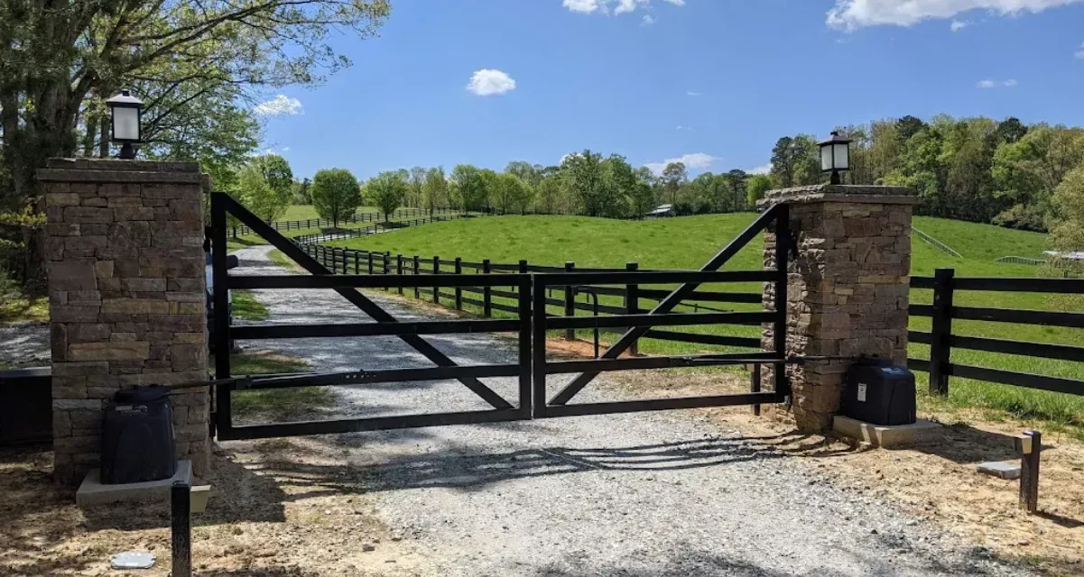 Stone gate posts with black wooden gates opening to a gravel driveway leading to a green field and trees under a blue sky.