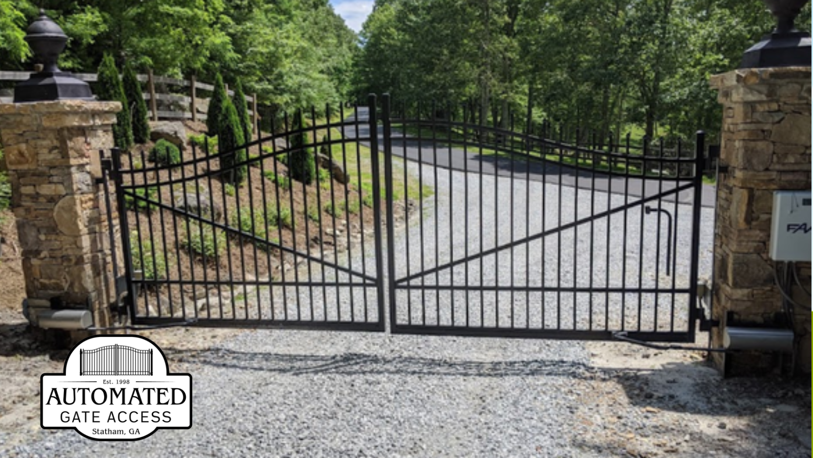 Black iron gate between stone pillars, leading to a gravel driveway surrounded by trees.