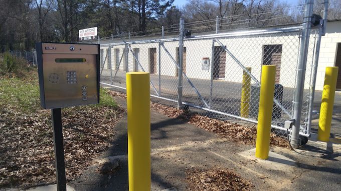 Metal keypad at a fenced storage facility entrance with yellow bollards.