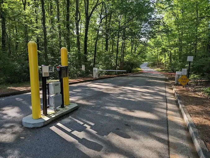 Entrance to a gated community. Yellow bollards and arm barrier block entry to a tree-lined asphalt road.