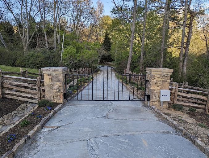 Driveway entrance with stone pillars, black metal gate, split-rail fencing, and surrounding trees.