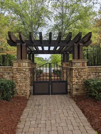 Stone and wood pergola with gate entrance; brick pathway, brown mulch, and green bushes.