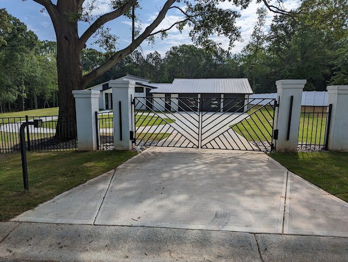 Black gate and driveway leading to a white house with a metal roof, surrounded by trees.