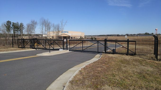 Black security gates open onto a paved road. Building and fence line in the background under blue sky.