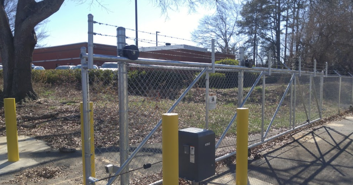 Chain link fence with barbed wire on top, a black gate opener box, and yellow bollards.