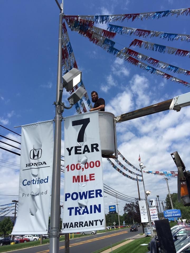 A man in a bucket lift working on a light pole at a Honda dealership, with banners and pennants.