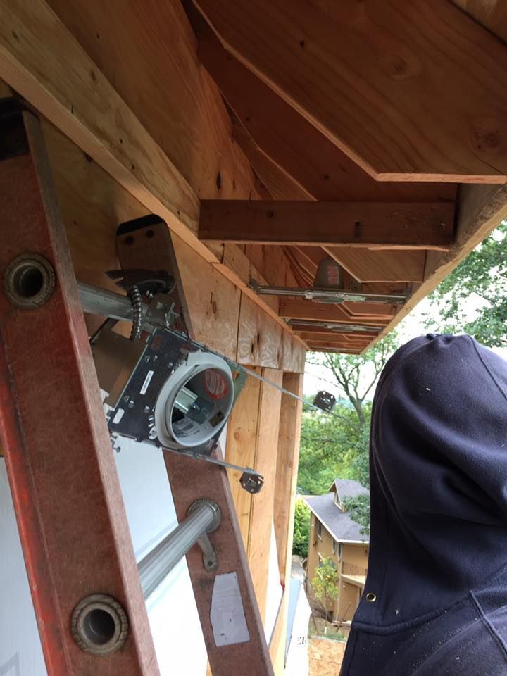 A person and ladder by a garage door mechanism being installed outside. Wooden frame is visible.
