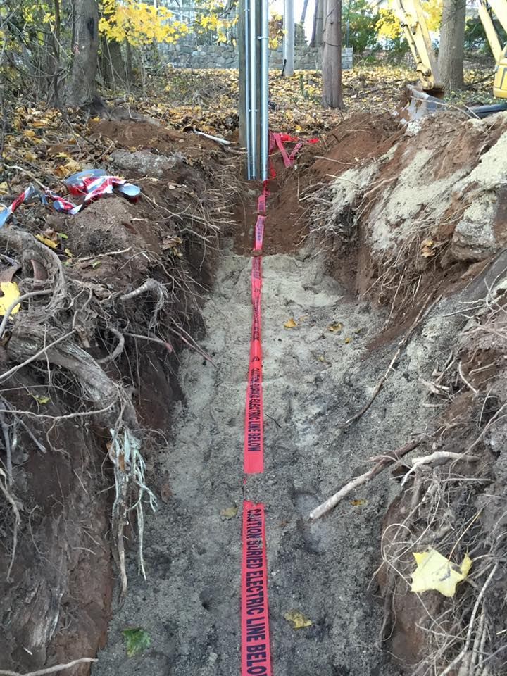 Trench dug in dirt, red tape warning of electric line below. Poles and trees in the background.