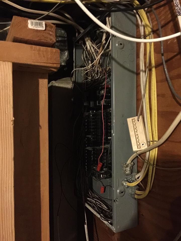 Electrical panel with exposed wires and breakers, mounted on a wooden wall in a basement setting.