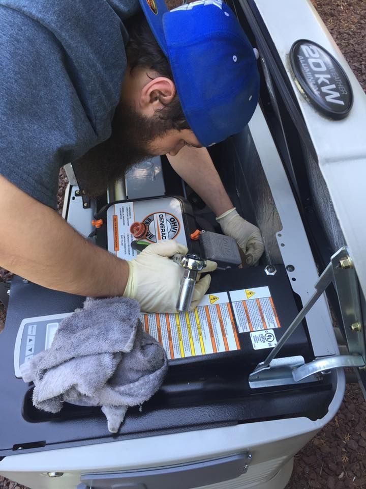 Mechanic wearing gloves working on a generator; they're holding a wrench and wearing a blue hat in an outdoor setting.