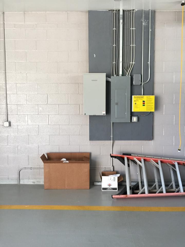 Electrical panel on gray painted wall with ladder, box, and conduit in a warehouse.