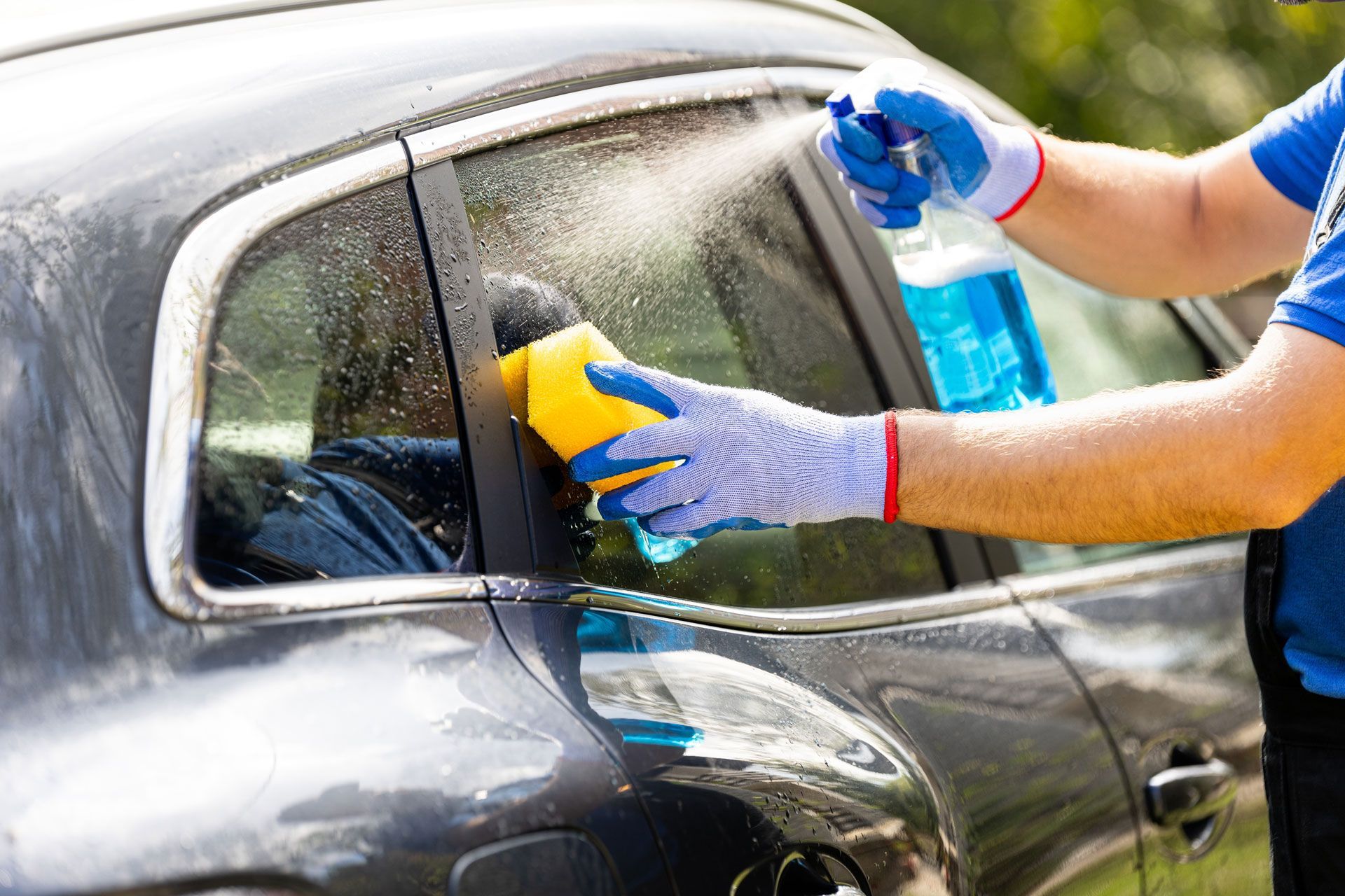 Person wearing gloves sprays and scrubs car window with a sponge.