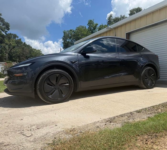 Black Tesla parked on a gravel driveway beside a garage under a partly cloudy sky
