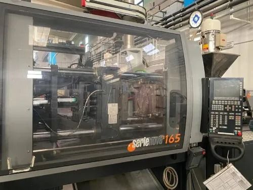 Macchina per stampaggio a iniezione industriale con pannello di controllo in uno stabilimento, vista attraverso una finestra di sicurezza.