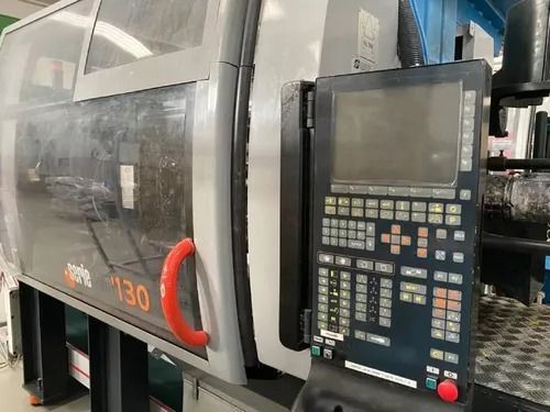 Pannello di controllo di una macchina industriale accanto a una pressa a iniezione grigia con maniglia rossa e schermo di visualizzazione.
