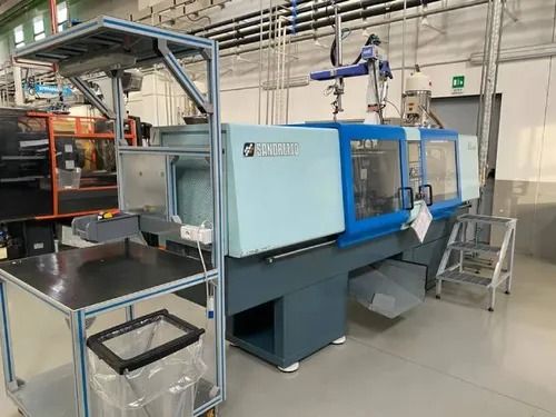 Grande pressa a iniezione industriale blu in una fabbrica con scalini in metallo e un banco da lavoro nelle vicinanze.