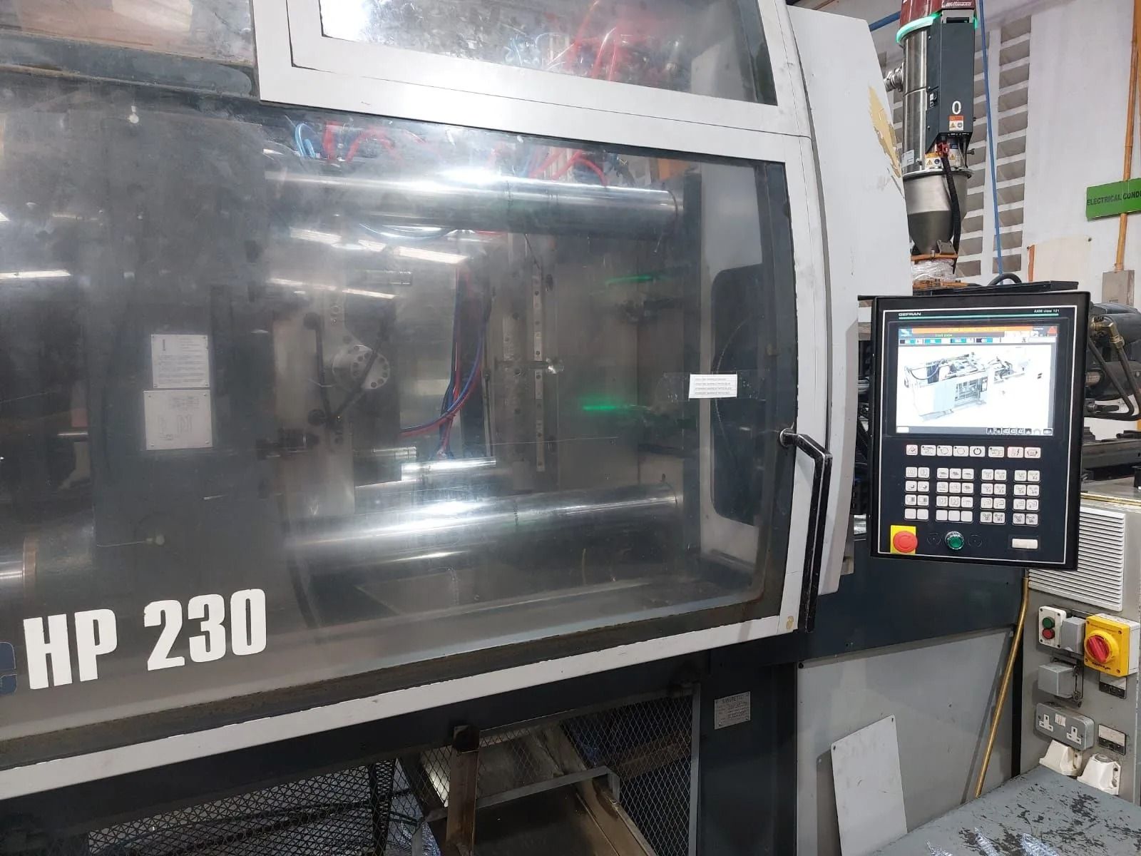 Pannello di controllo di una macchina industriale accanto a una grande macchina HP 230 grigia in un ambiente di fabbrica.