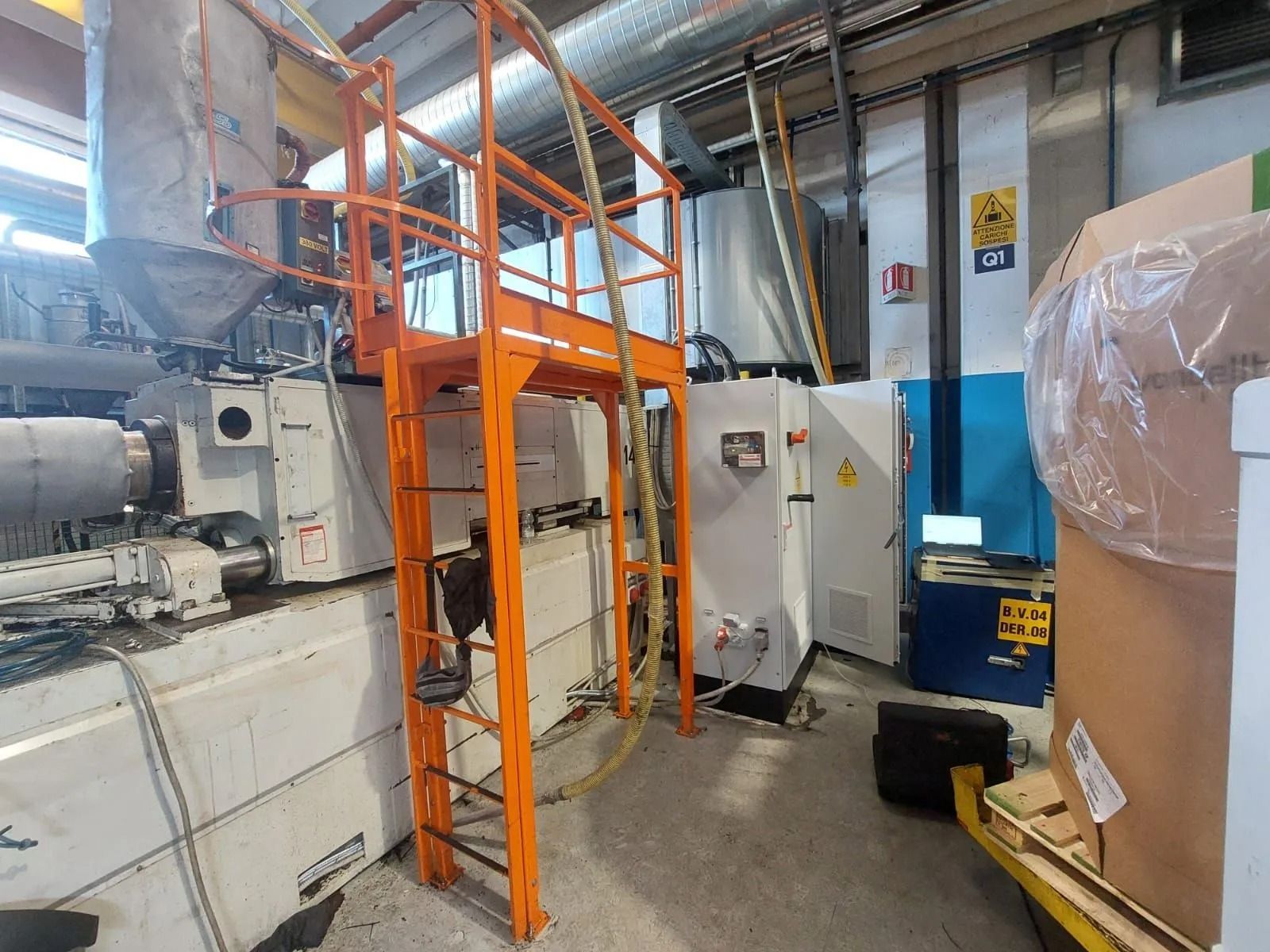 Un ascensore industriale arancione all'interno di una fabbrica, accanto a grandi macchinari e attrezzature imballate.