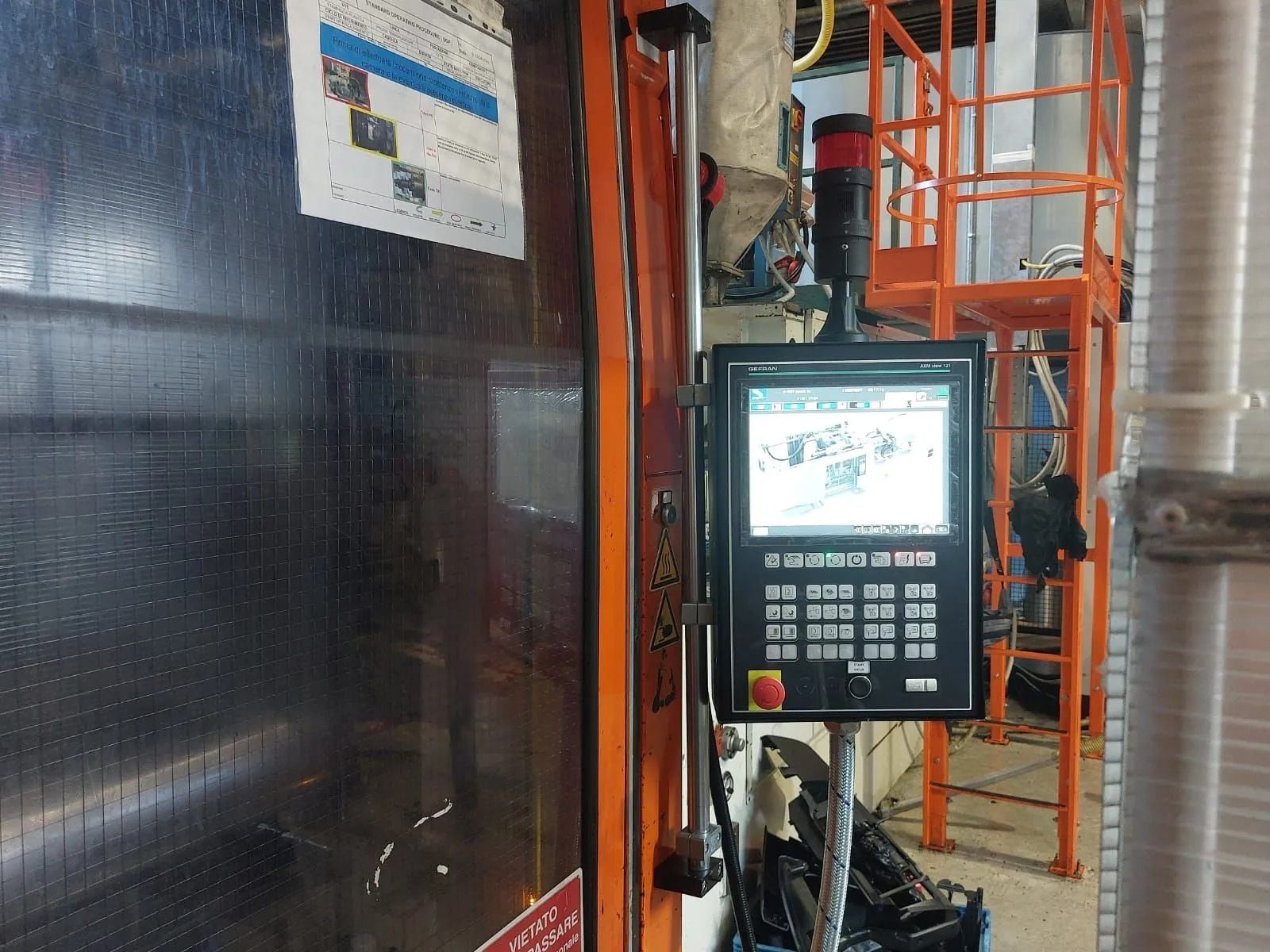 Pannello di controllo industriale montato accanto a un grande macchinario nero in una fabbrica, con ringhiere di sicurezza arancioni sullo sfondo.