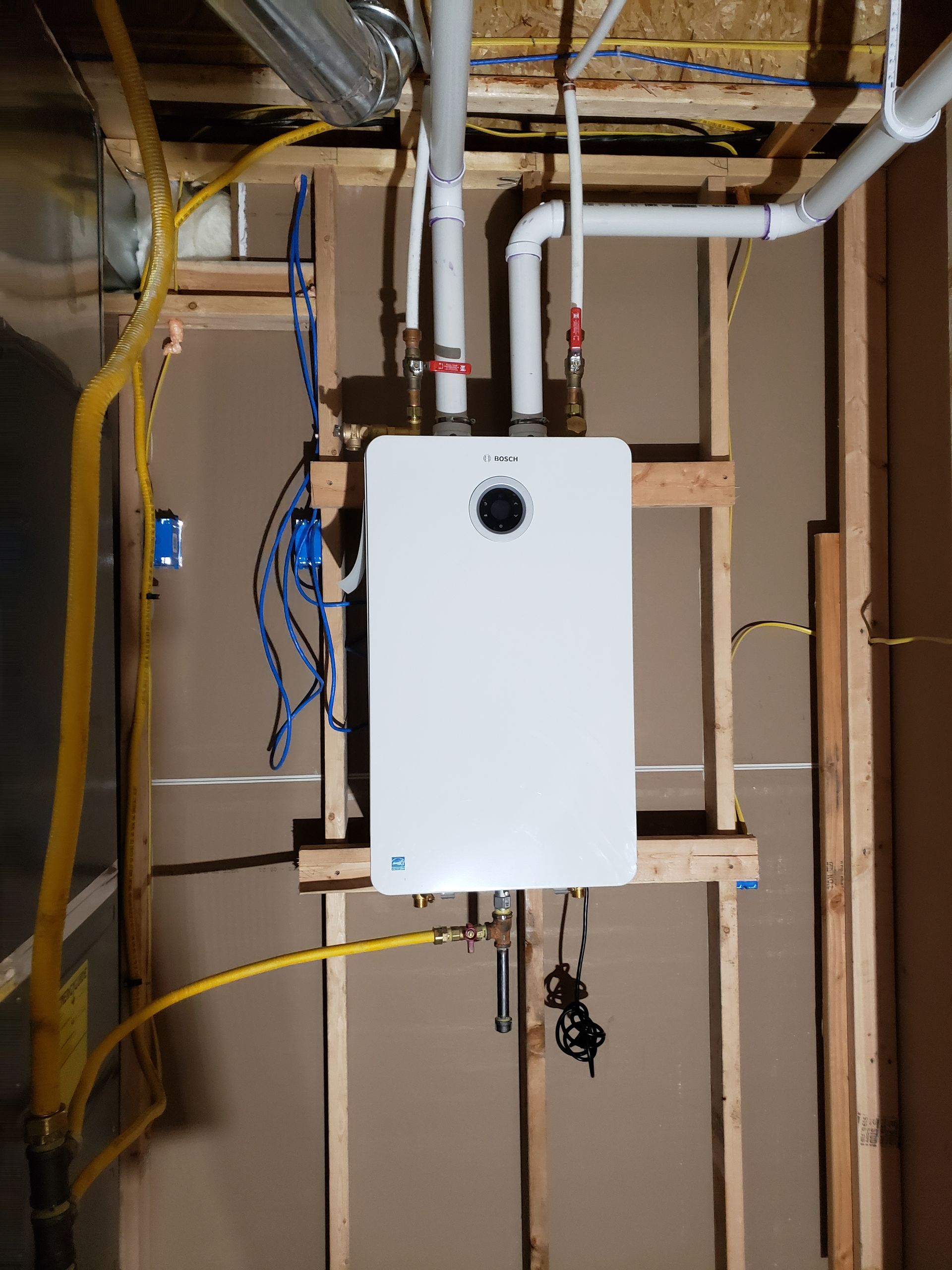 White wall-mounted boiler with exposed pipes and wiring in an unfinished basement utility area