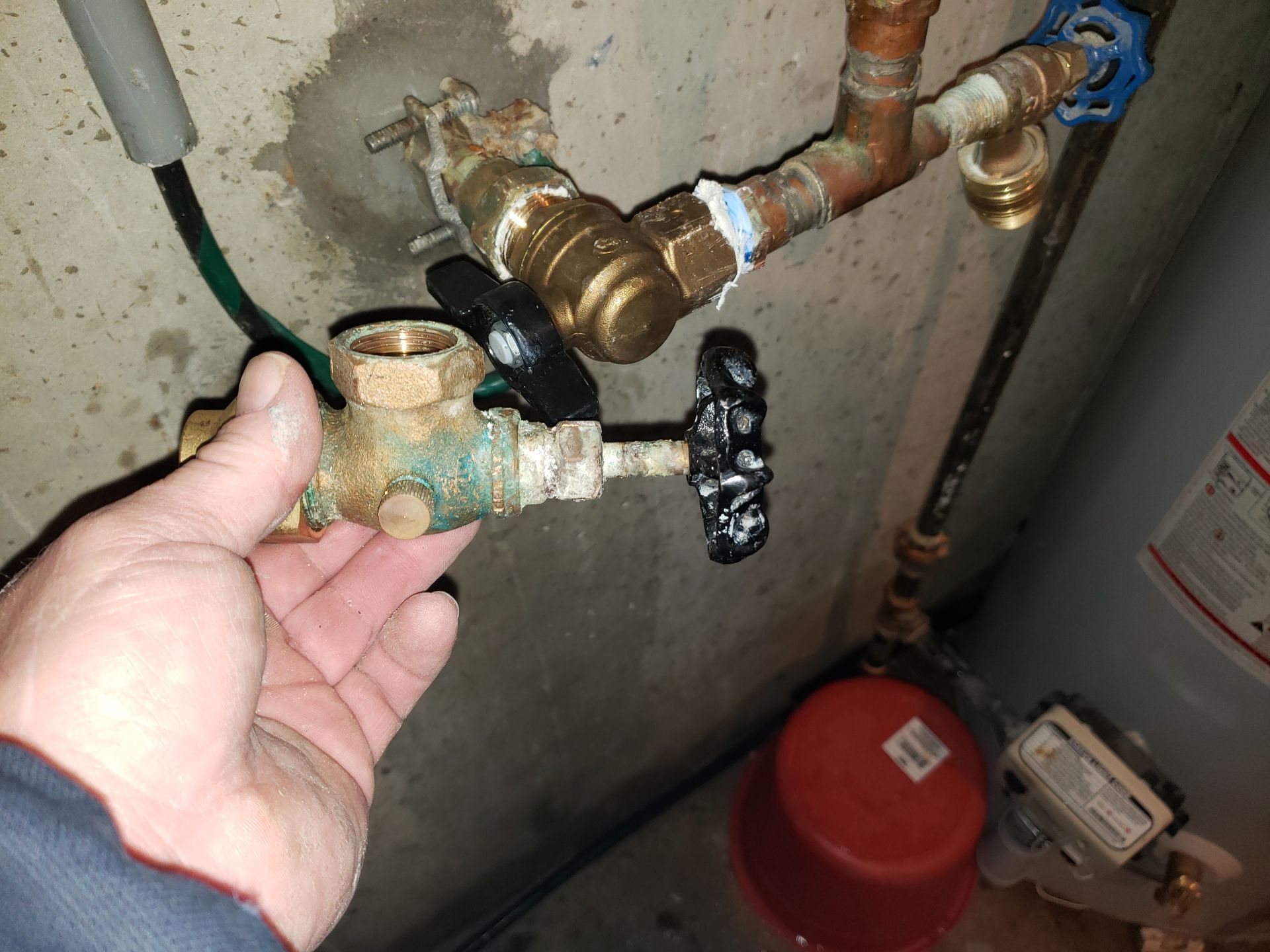 Hand holding a green valve on exposed plumbing pipes beside a water heater.