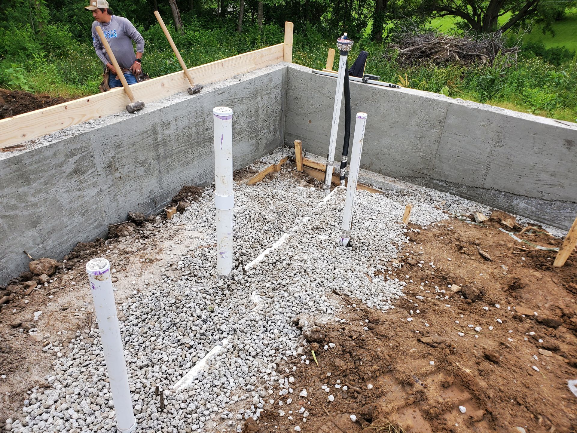 Concrete foundation walls with gravel and white PVC pipes in an excavation; a worker stands at the edge.