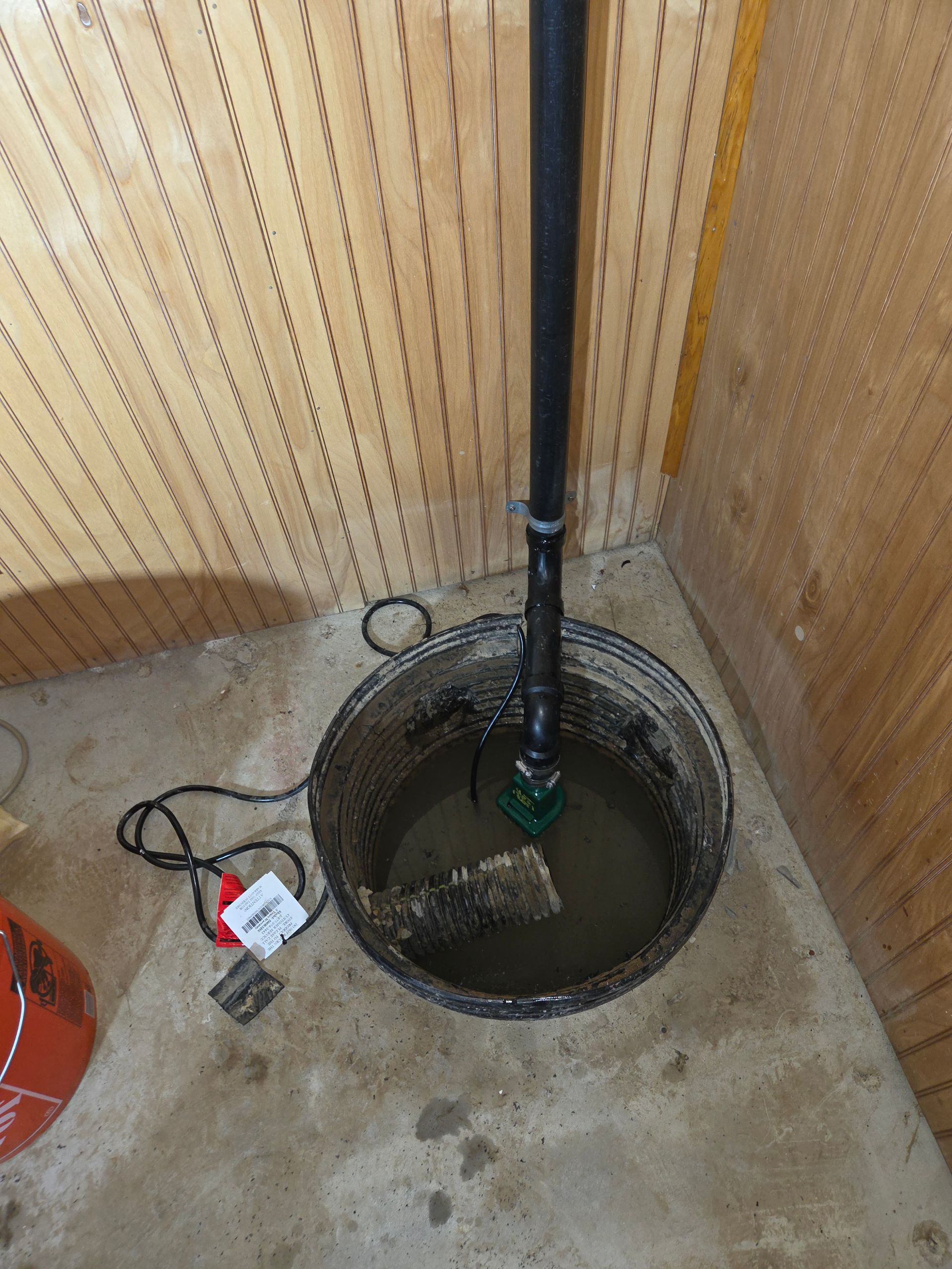 Open sump pit with black pump pipe in a concrete utility room; orange bucket and cords nearby