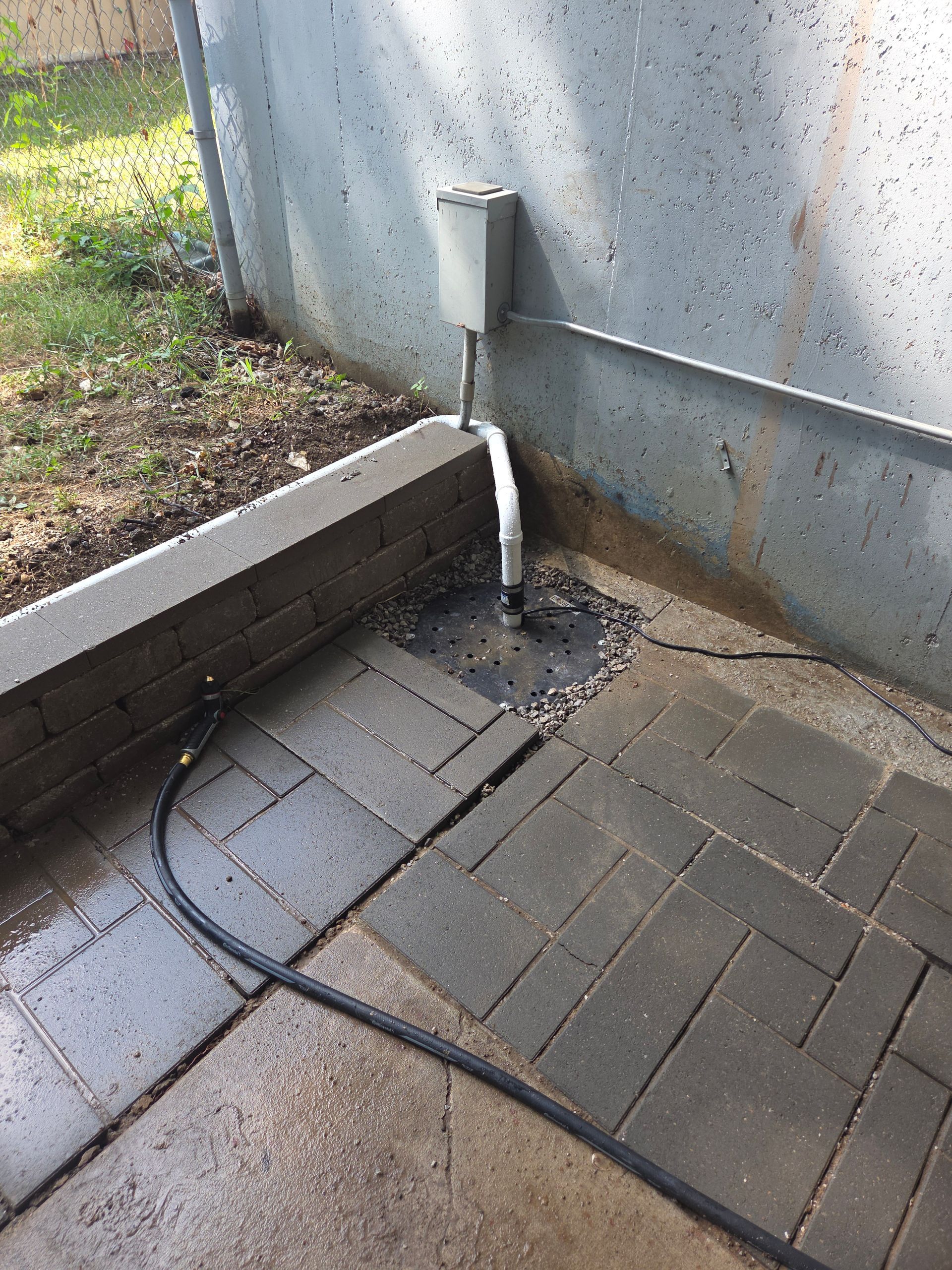 Small downspout drains onto wet patio pavers beside a concrete wall and low brick border.