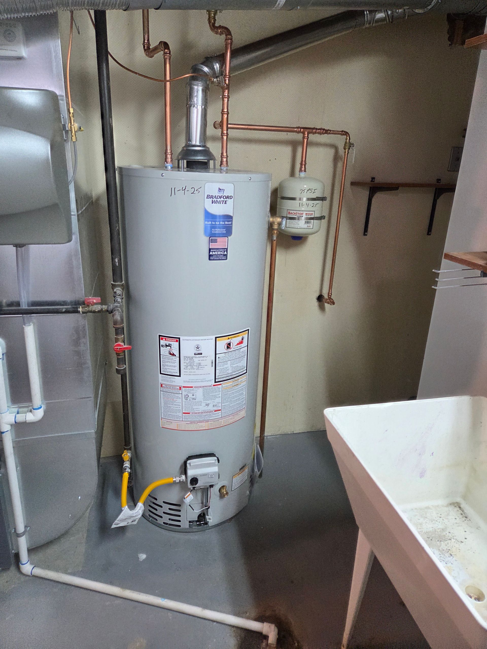 Water heater in a utility room with exposed pipes and a nearby sink.