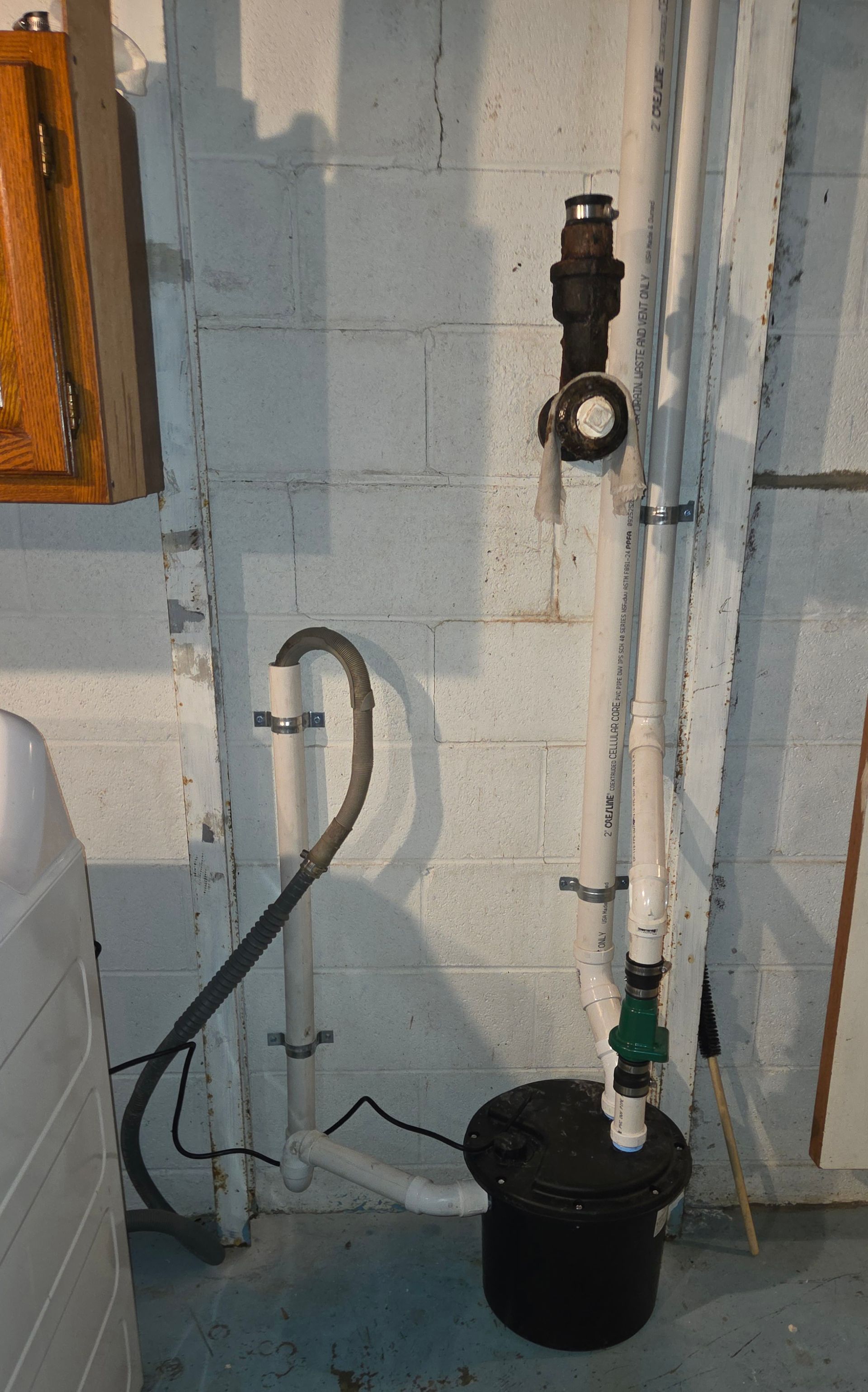 Utility pipes and a black bucket in a small basement corner against a white brick wall.
