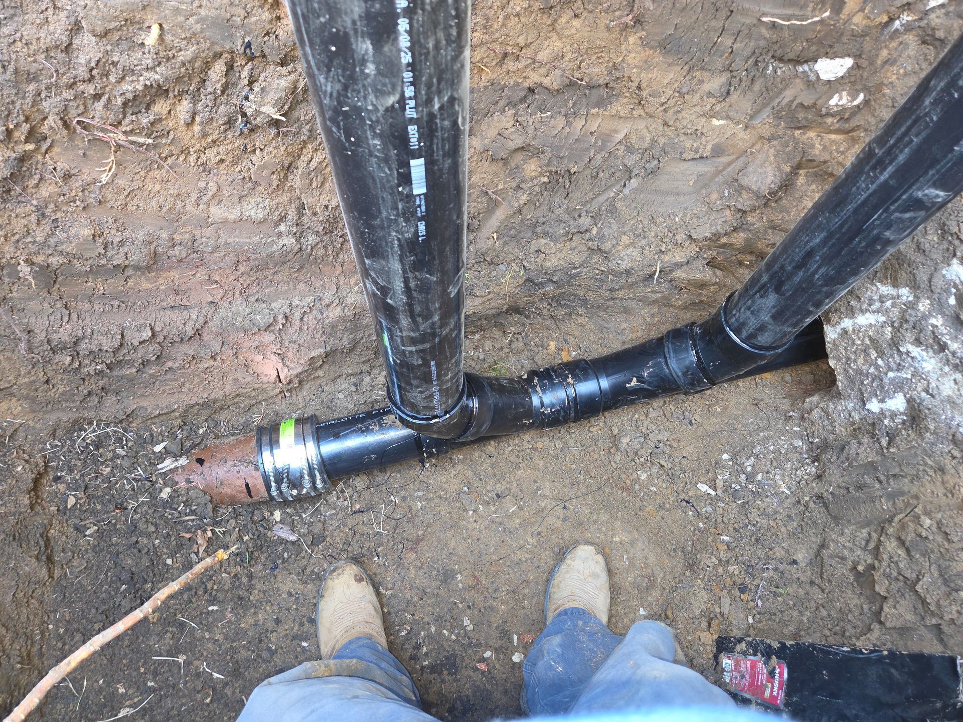 Black pipes joined in a trench beside workers’ boots and dirt ground