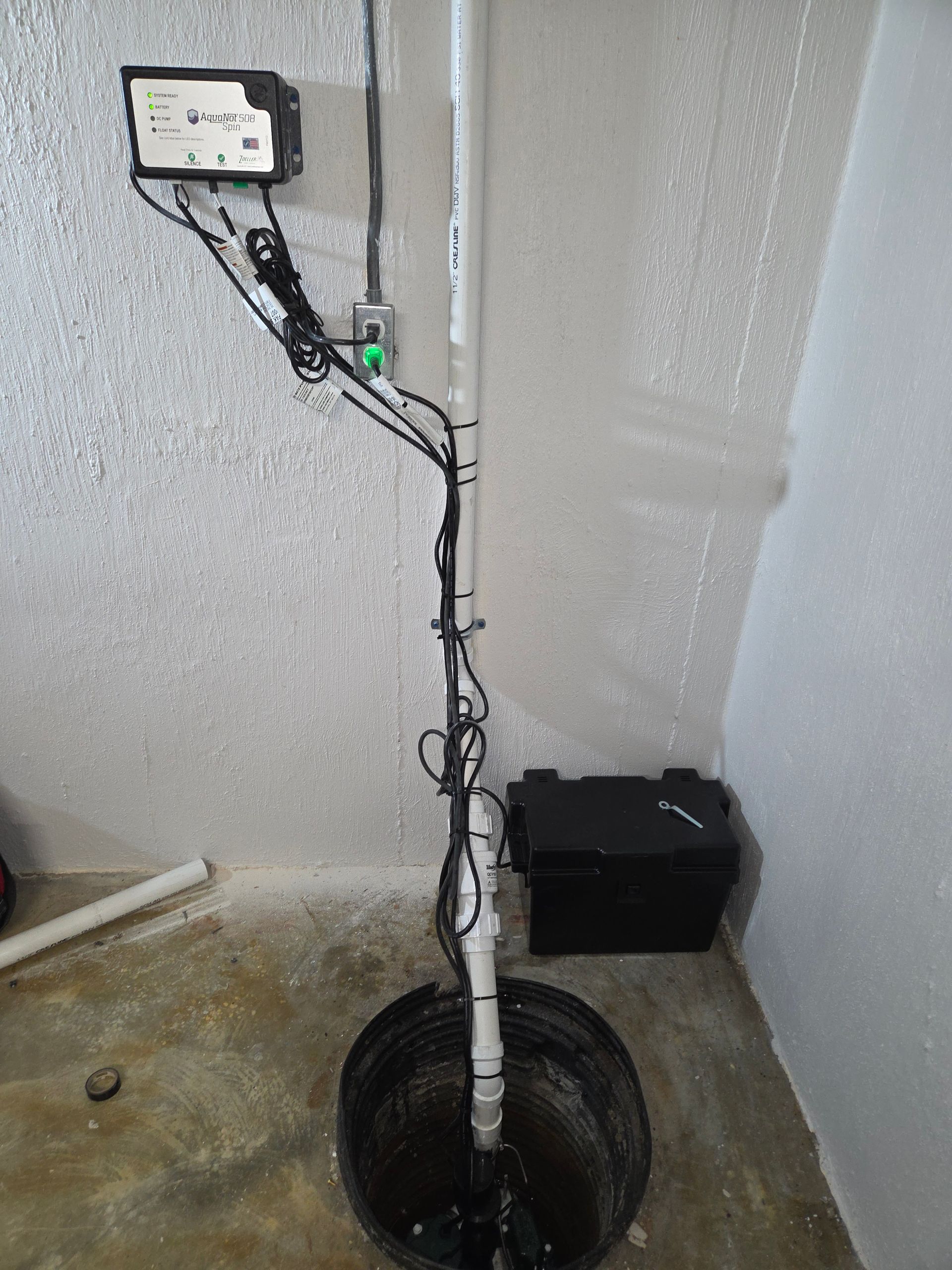 Sump pump pit with vertical pipe and wiring in a utility room corner, next to a black storage bin.