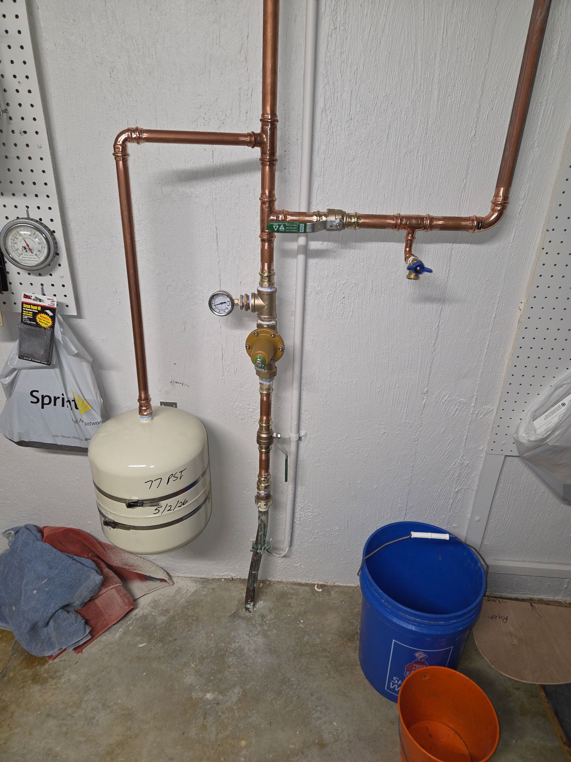 Basement utility wall with copper pipes, pressure tank, and blue and orange buckets on the floor